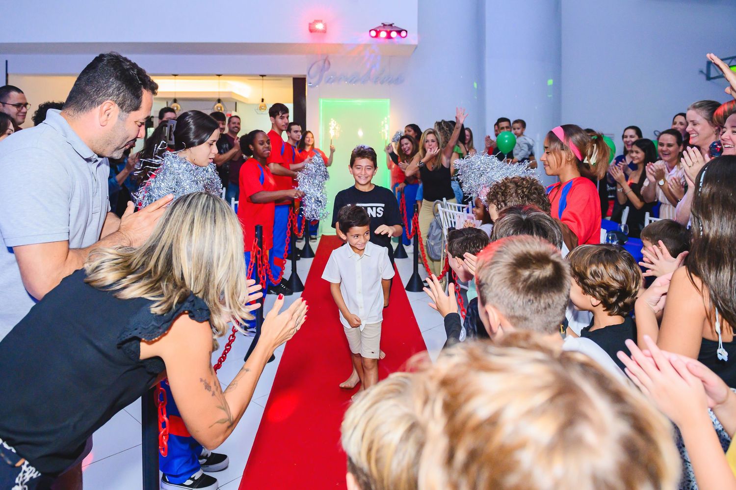 Excited children gather around a red carpet at an indoor event space while adults photograph the special occasion.