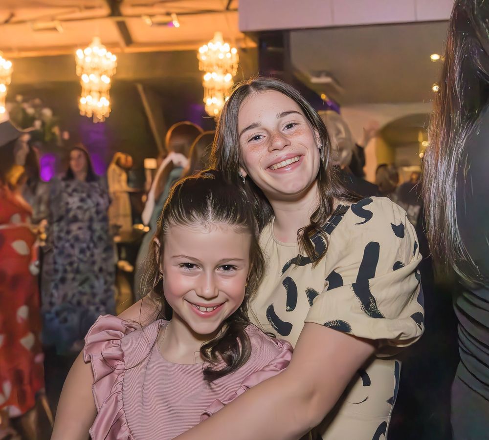 Two partygoers in pink and beige outfits smile together at a festive indoor event with decorative lighting and guests.