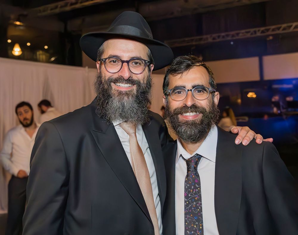 Two bearded men in suits and glasses smile together at a formal event, one wearing a black hat.