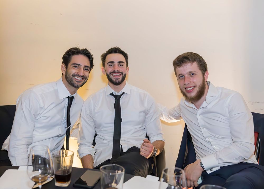 Three friends in white dress shirts and ties smile while sitting together at a restaurant table with wine glasses.