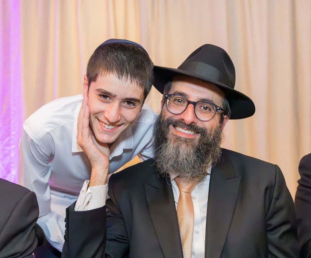 Two people posing together at an event - one wearing a white dress shirt and one in traditional Orthodox Jewish attire.