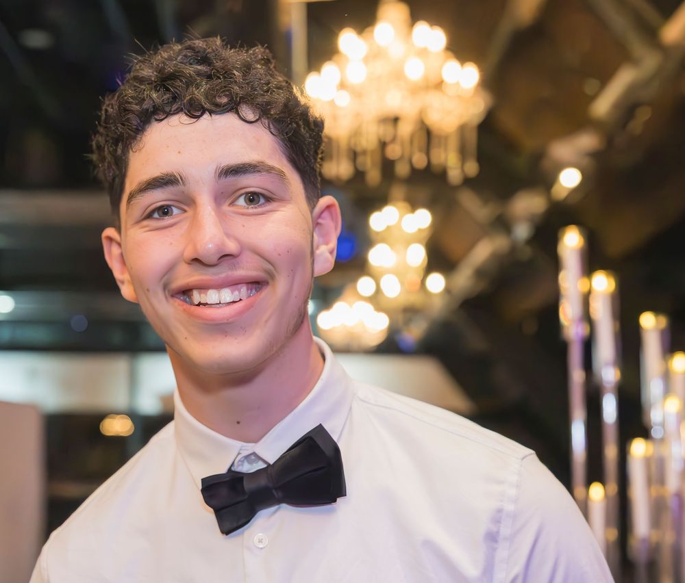 A waiter in black bow tie smiles warmly while serving at an upscale event with crystal chandeliers.