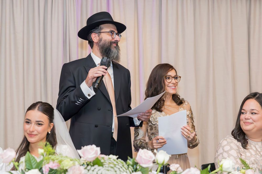 People gather at an elegant event with flowers while someone in a black suit and hat gives a speech at a table.