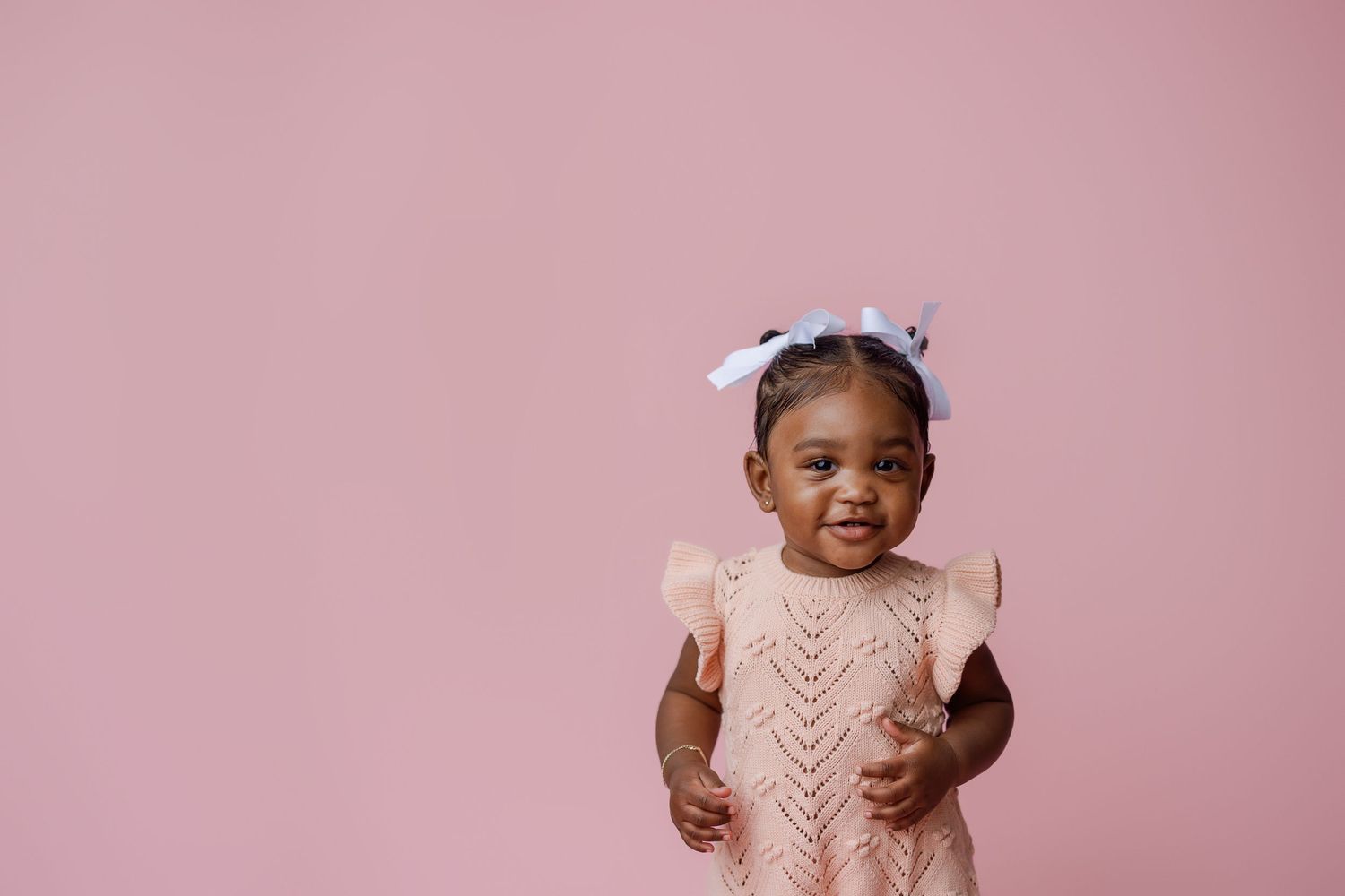 A smiling young child wearing a light pink knitted sweater dress with ruffled sleeves against a pink background.
