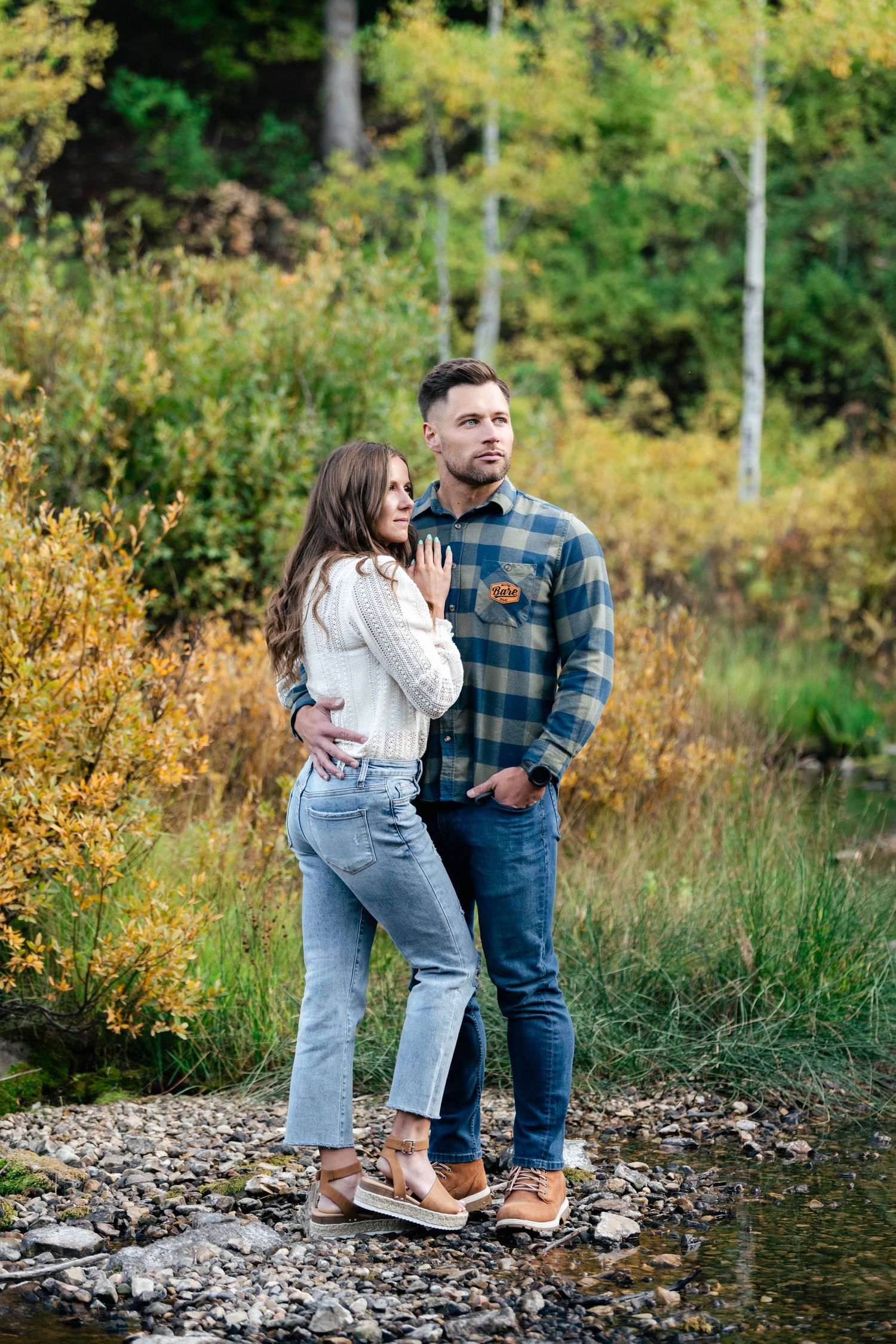 A couple shares tender moments near a rocky shoreline surrounded by autumn foliage in a natural outdoor setting.