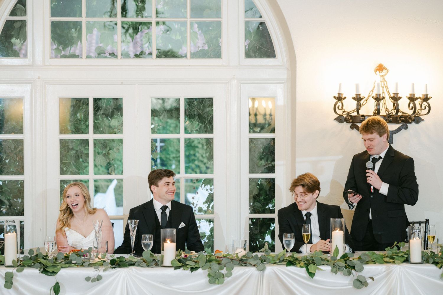 Elegant wedding reception table at a venue with arched windows and chandelier featuring floral centerpieces and formal place settings.