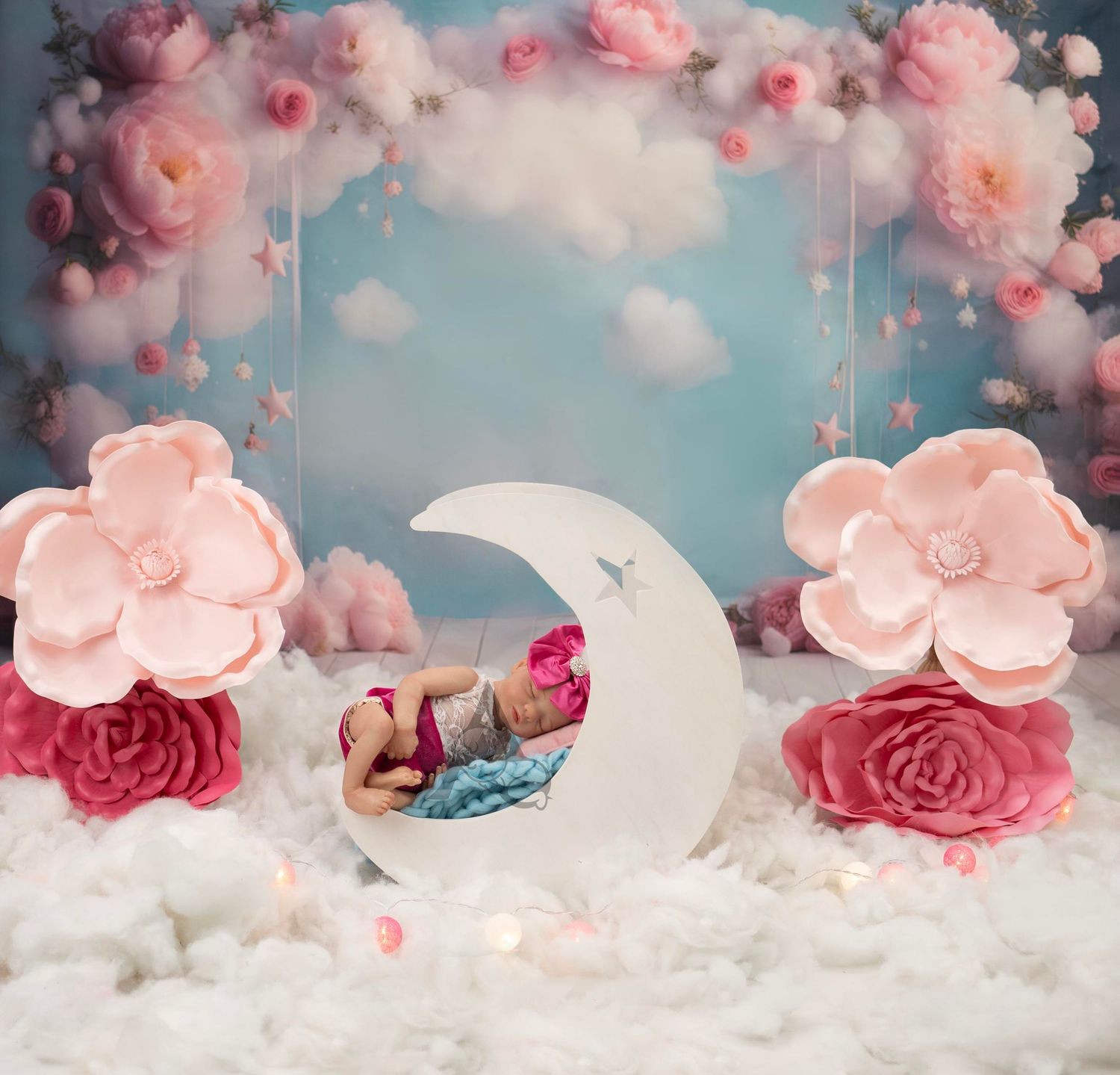 Whimsical baby photography setup with crescent moon prop amid pink roses and clouds.