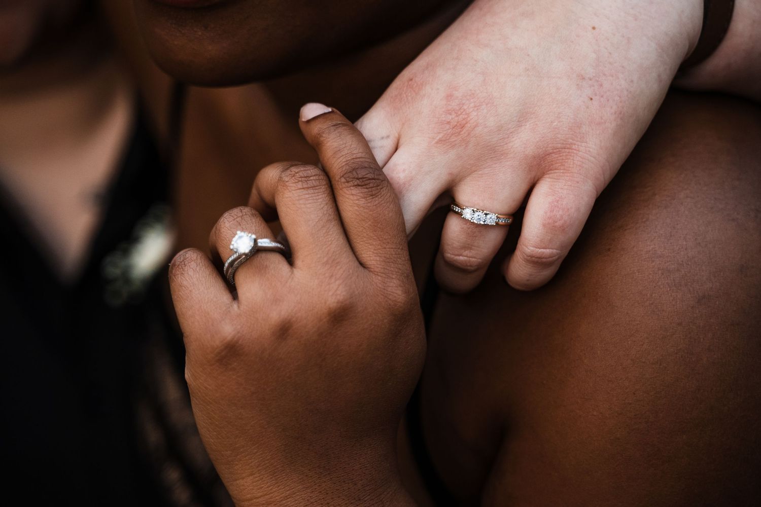 Two hands wearing engagement rings touch in a romantic close-up moment against a dark background.