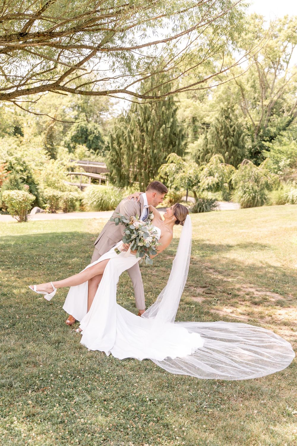 Romantic wedding scene with couple in light beige and white sharing a dip kiss under trees on sunny lawn.