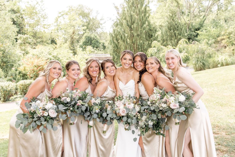 Bridesmaids in champagne dresses pose together with bouquets for a joyful group photo.