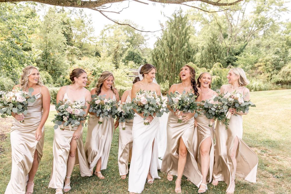 Bridal party in champagne silk dresses laugh together while walking through a sunny garden setting.