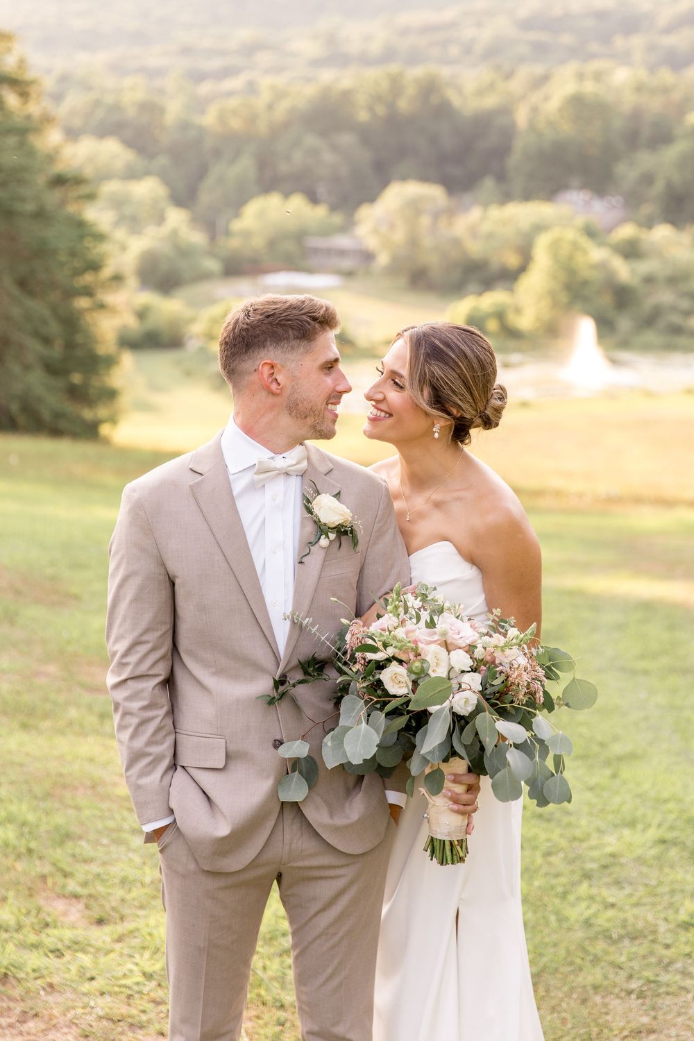 Newlywed couple poses outdoors wearing light colored wedding attire with a lush green natural background and bouquet.