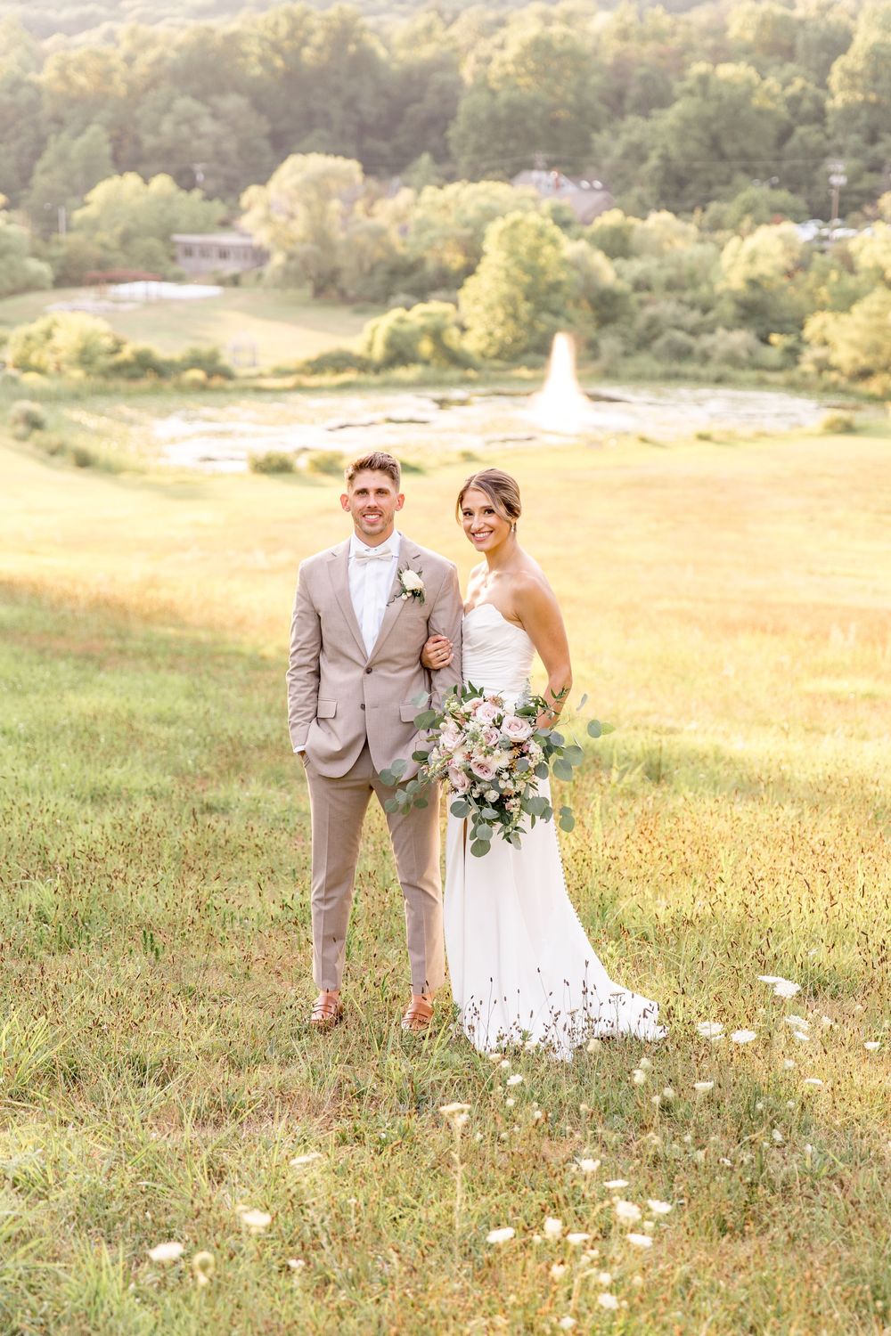 A couple stands in a golden meadow for their wedding portrait with a fountain and trees visible in the background.