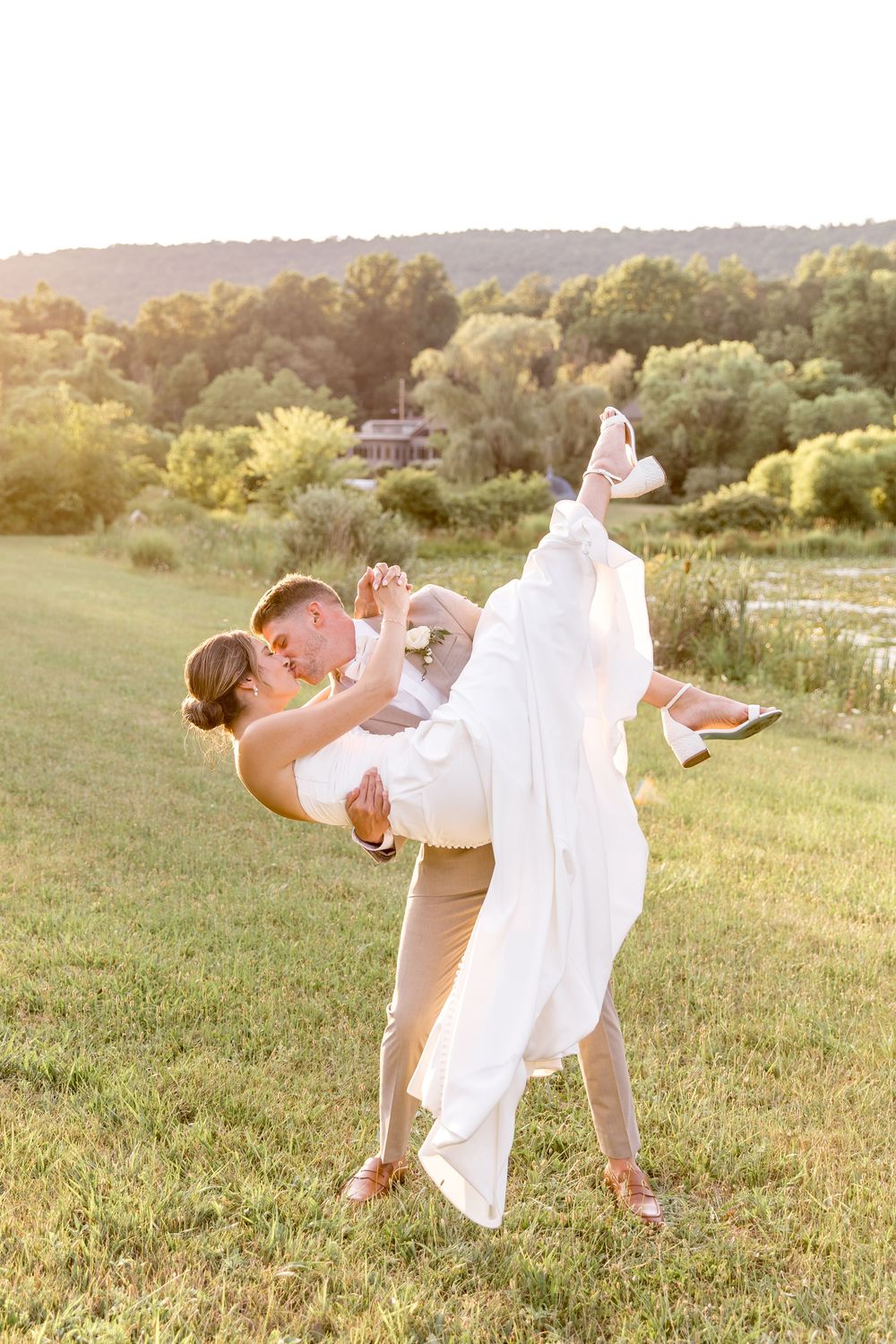 A romantic wedding couple embraces in a sunlit meadow during golden hour with rolling hills in the background.