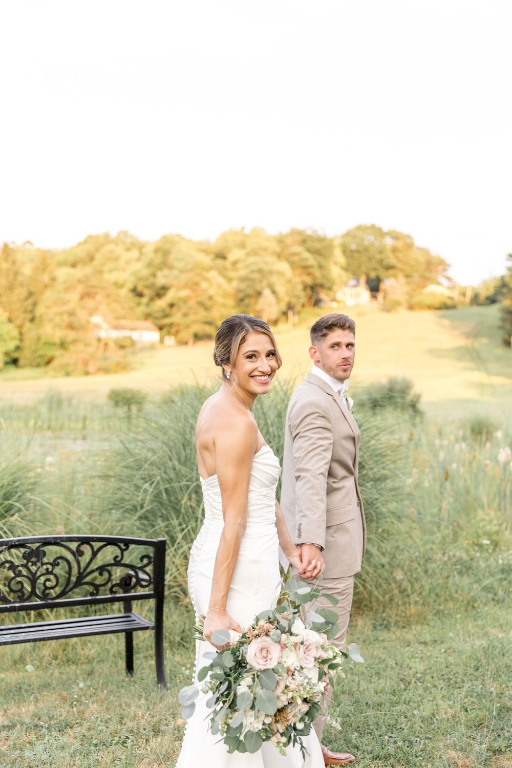 Bride holds bouquet while couple poses near decorative bench in golden evening light.