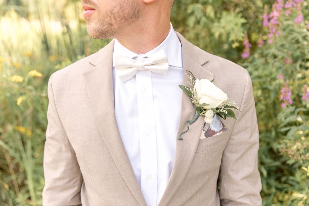 Close up of groom's formal attire featuring a tan suit jacket, white bowtie and rose boutonniere.