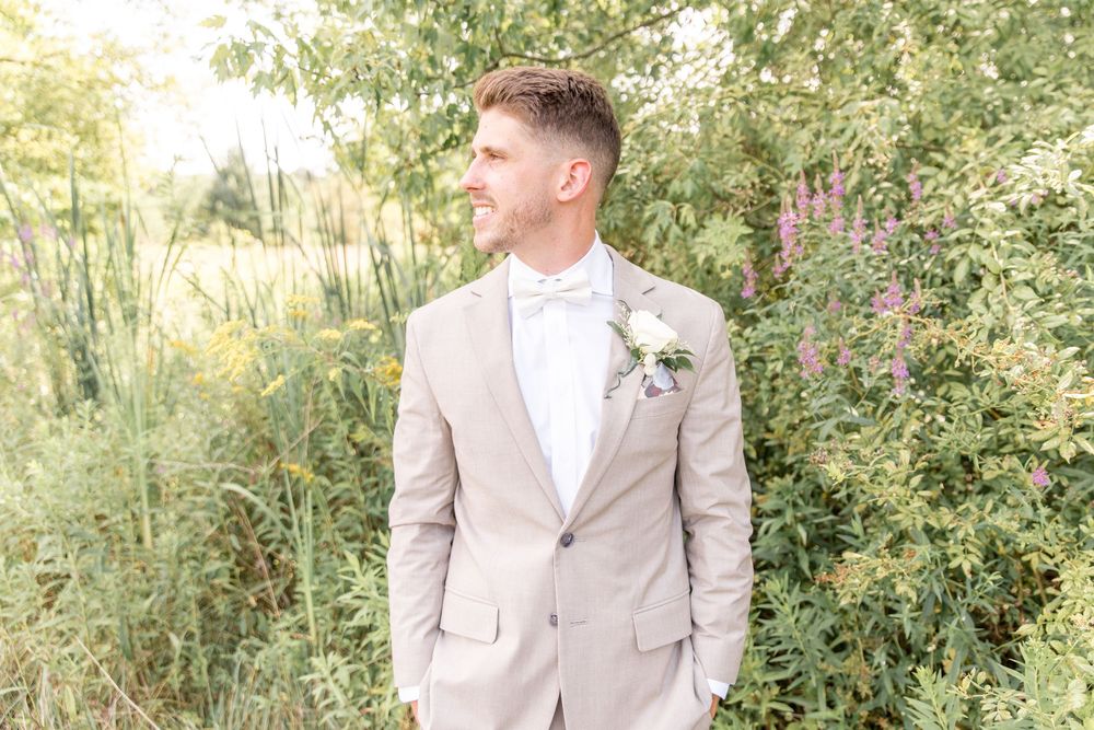Groom in light beige suit with white boutonniere stands against lush garden backdrop during outdoor wedding.