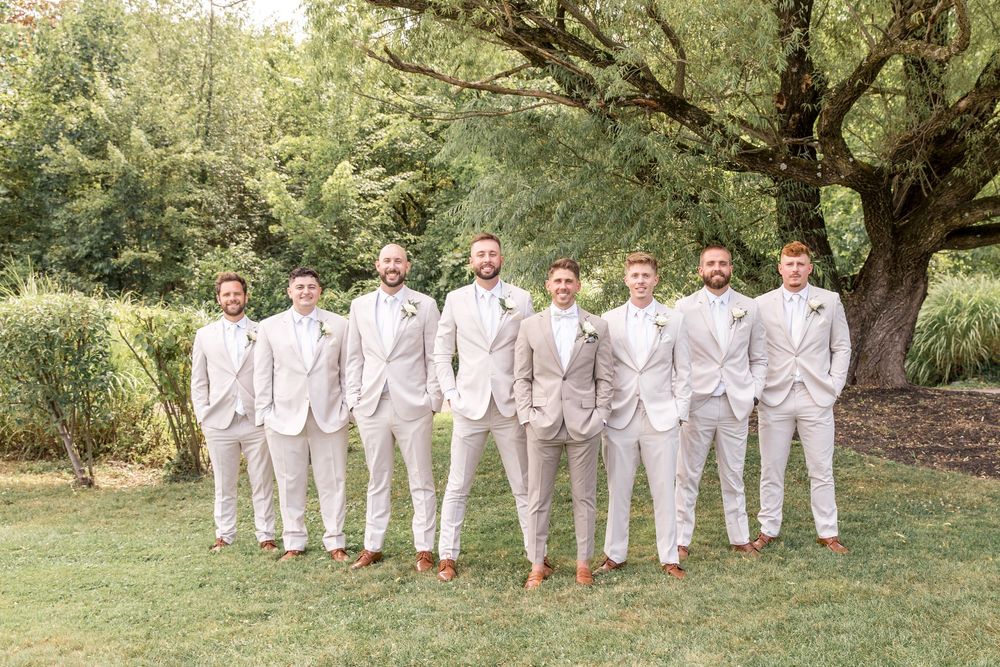 Groomsmen in light gray suits and brown shoes lined up outdoors for formal wedding photo.