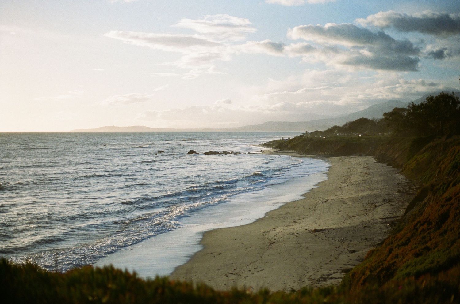 Scenic coastal view of waves rolling onto a curving sandy beach beneath cloudy skies at sunset.