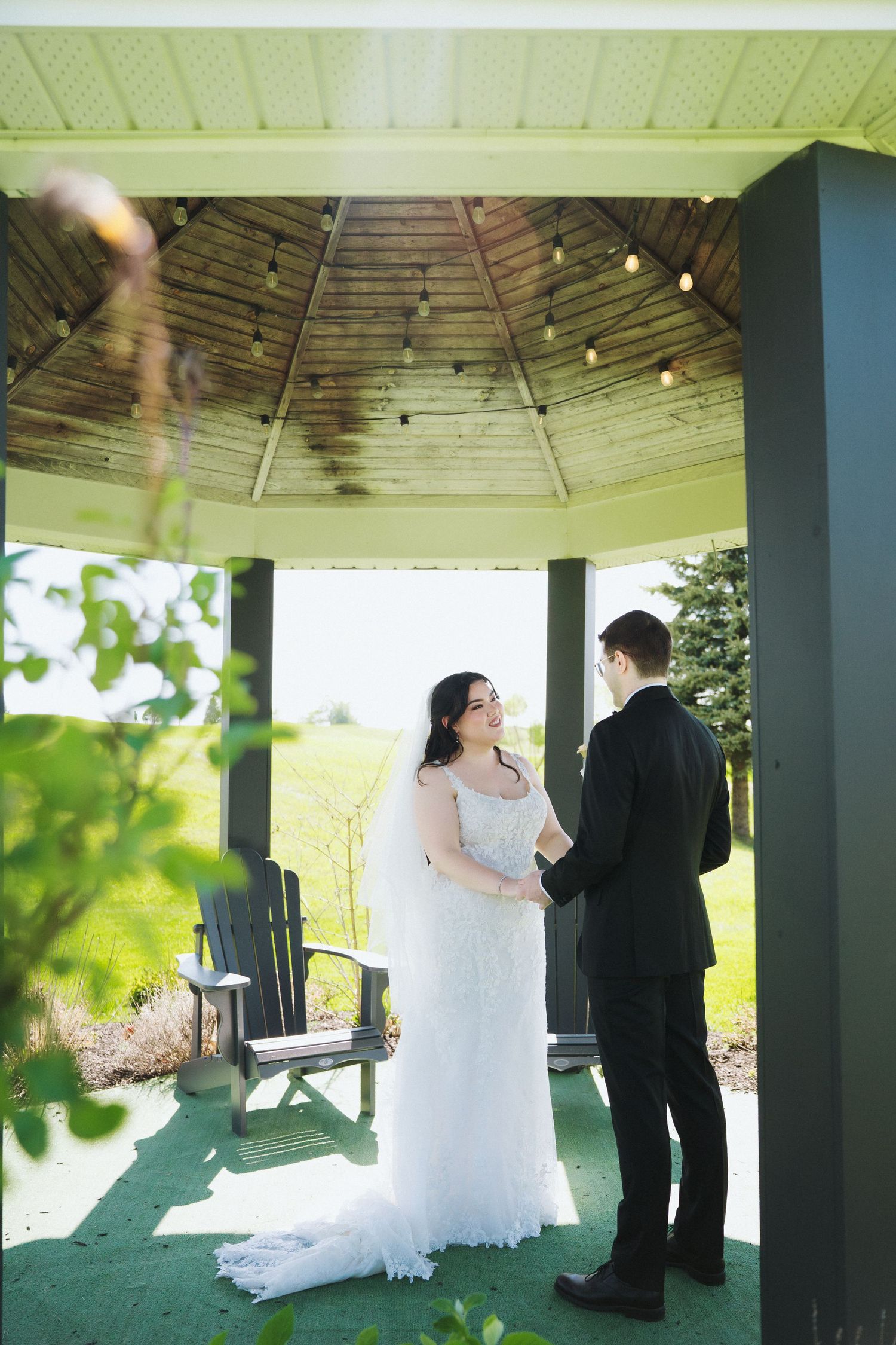 Wedding couple exchange vows under wooden gazebo with string lights.