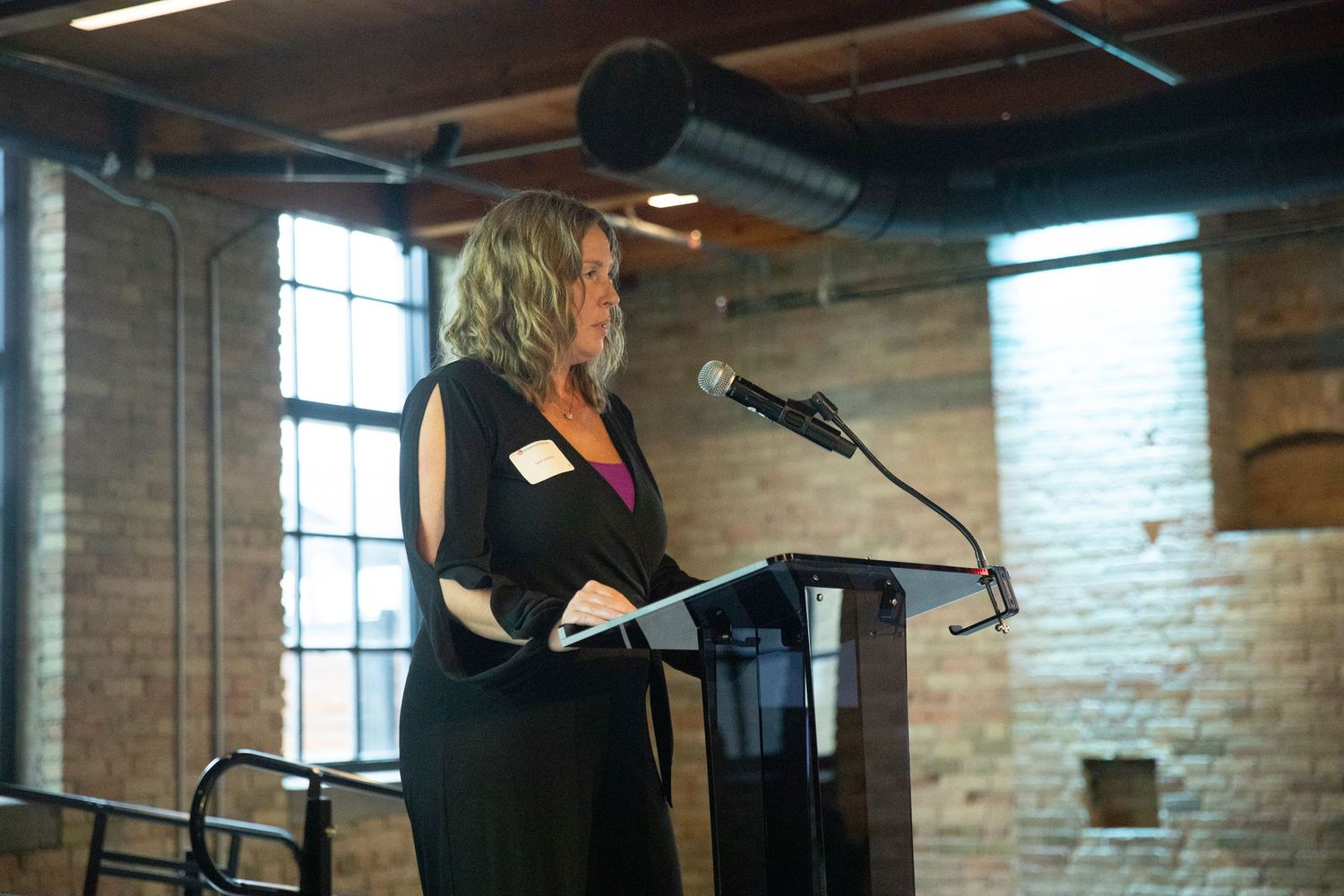A speaker stands at a podium in an industrial-style room with exposed brick walls and ductwork.