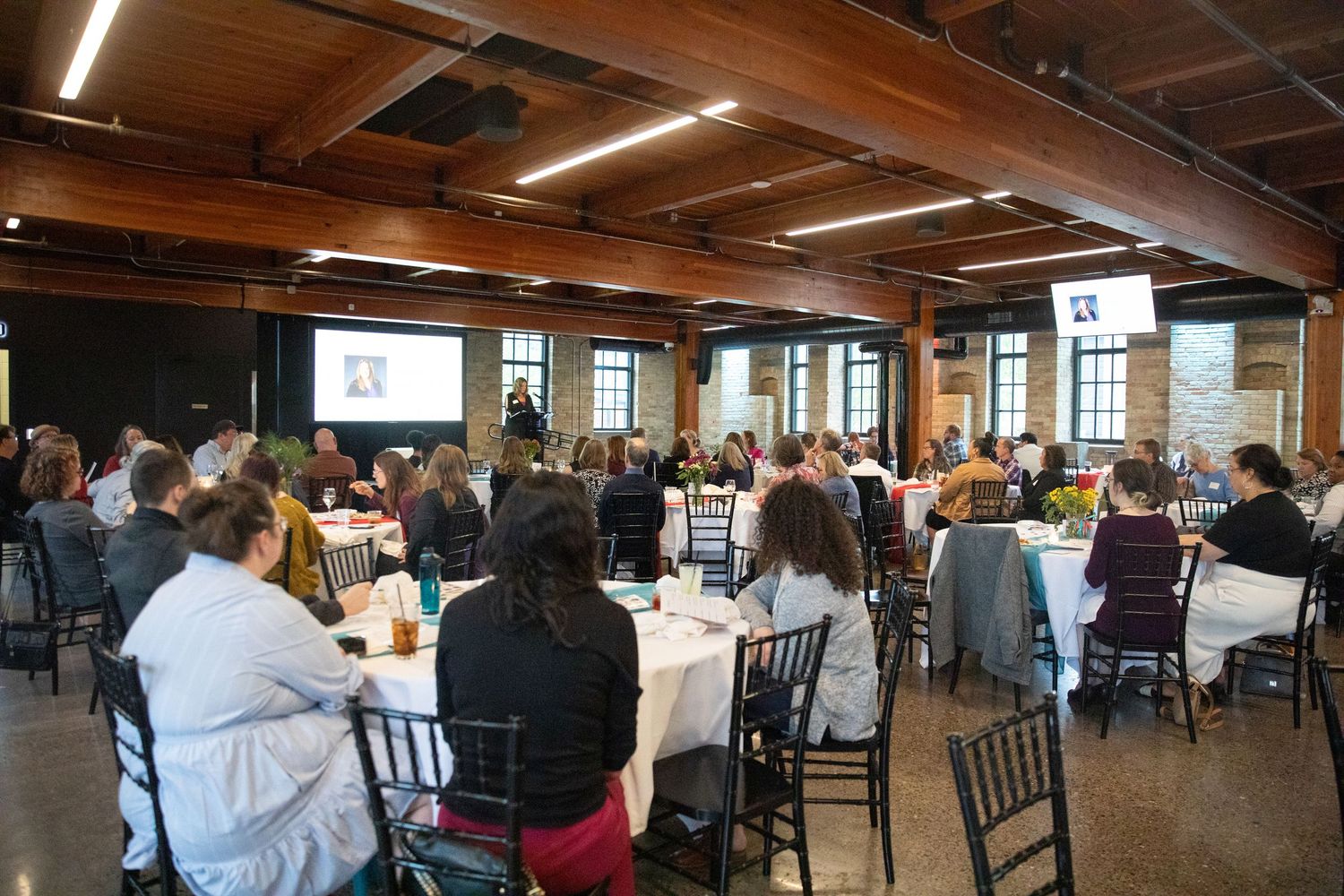 A large group of people attend a conference in a rustic event space with exposed ceiling beams.