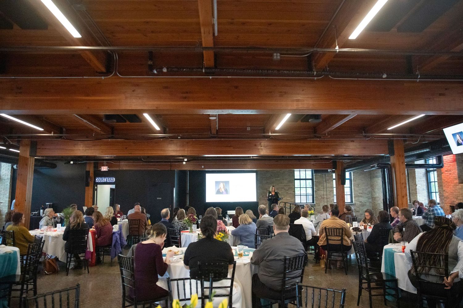 A large group of attendees seated at round tables in an industrial-style venue with exposed wooden beams and lighting.