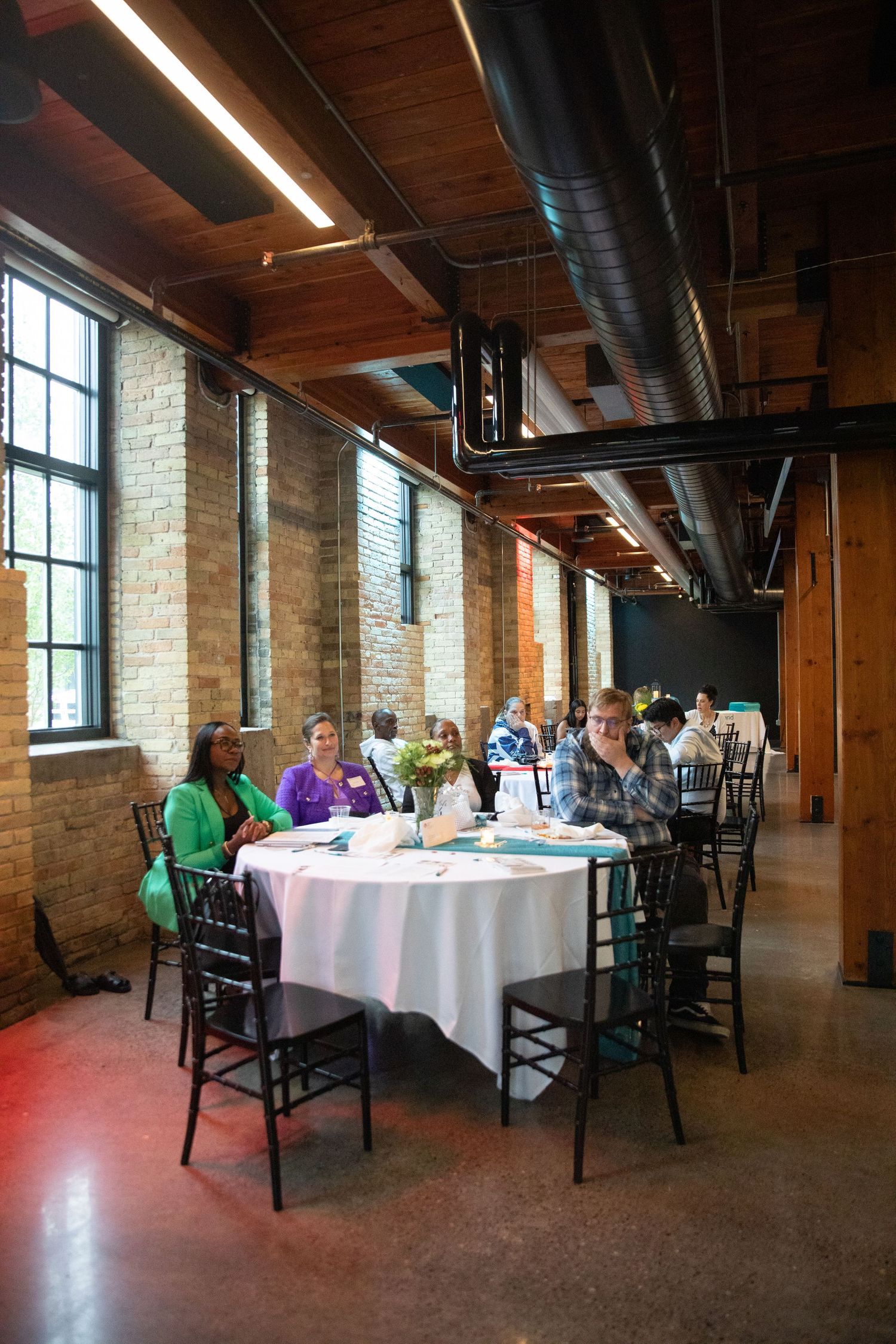 Large round tables with white tablecloths set up in an industrial venue with exposed brick walls and high windows.