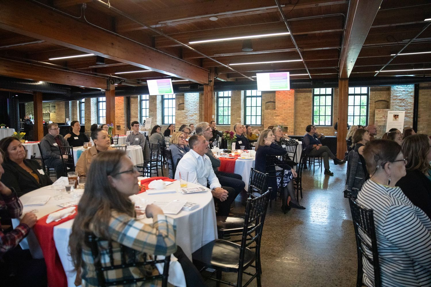 A crowded business luncheon in an industrial-style venue with exposed brick walls and wooden beams.
