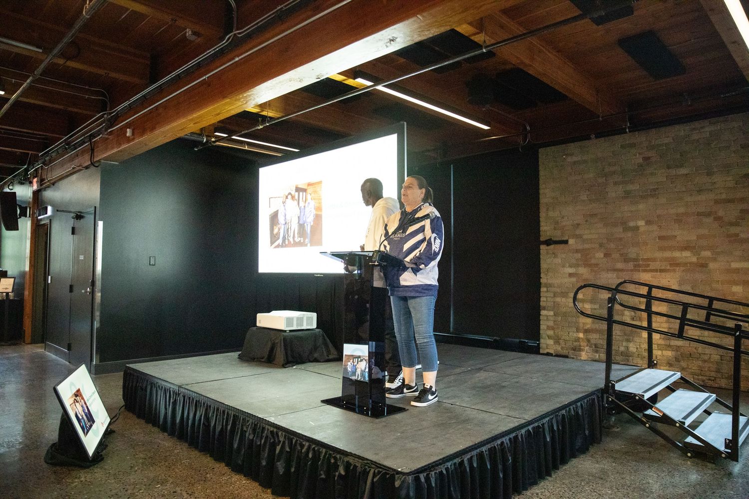 Two presenters stand on a black stage giving a presentation in an industrial-style venue.