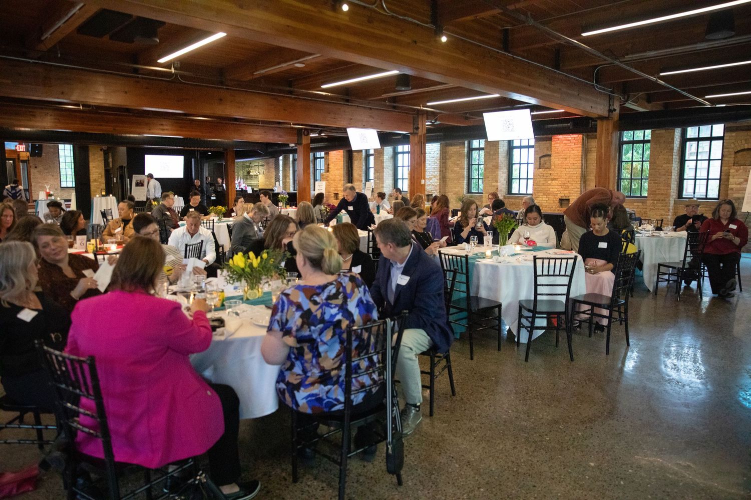 Conference attendees gather at round tables in a rustic venue with exposed ceiling beams and large windows.