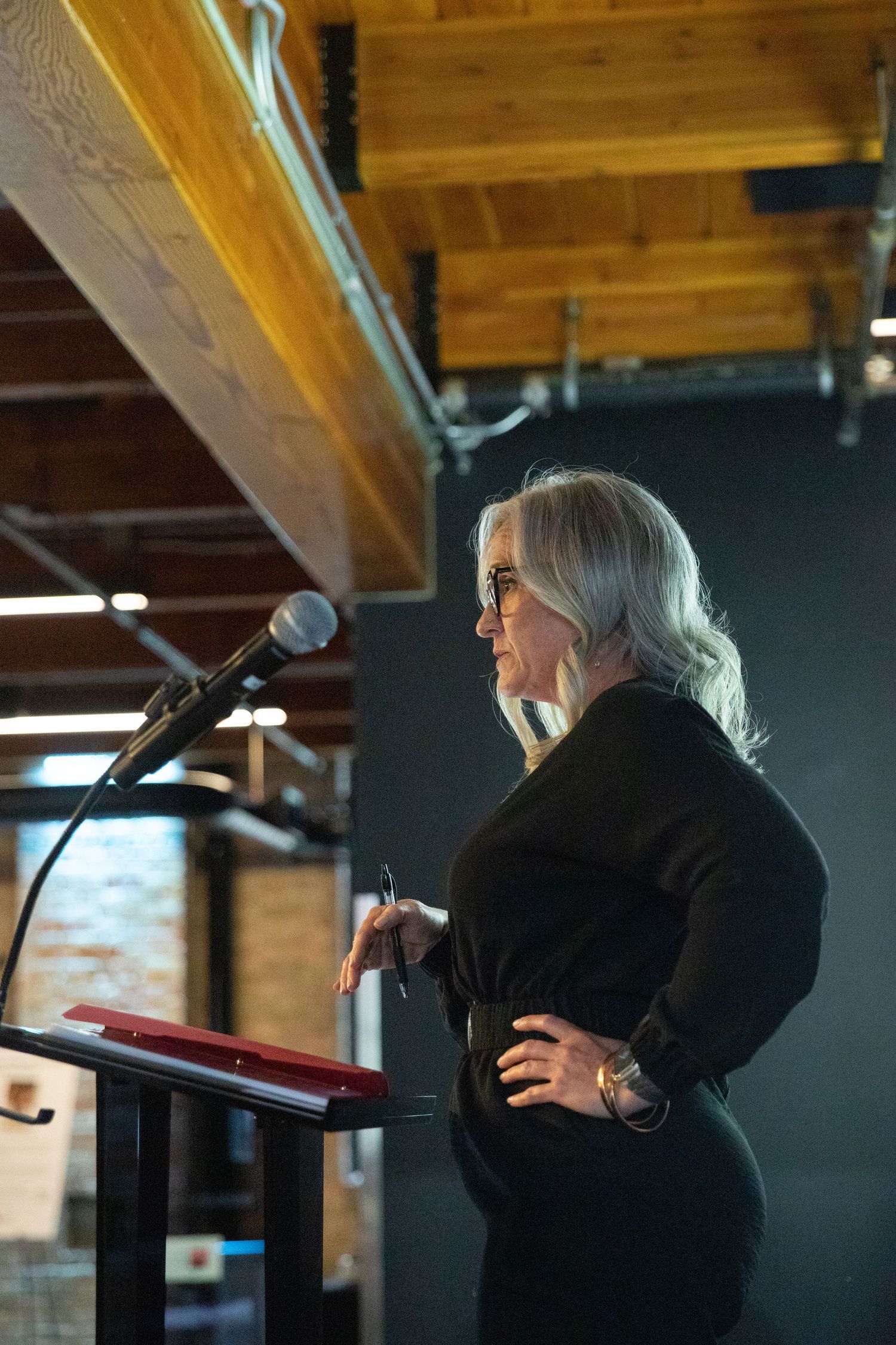 A speaker in black attire stands at a podium with a microphone under exposed wooden ceiling beams.