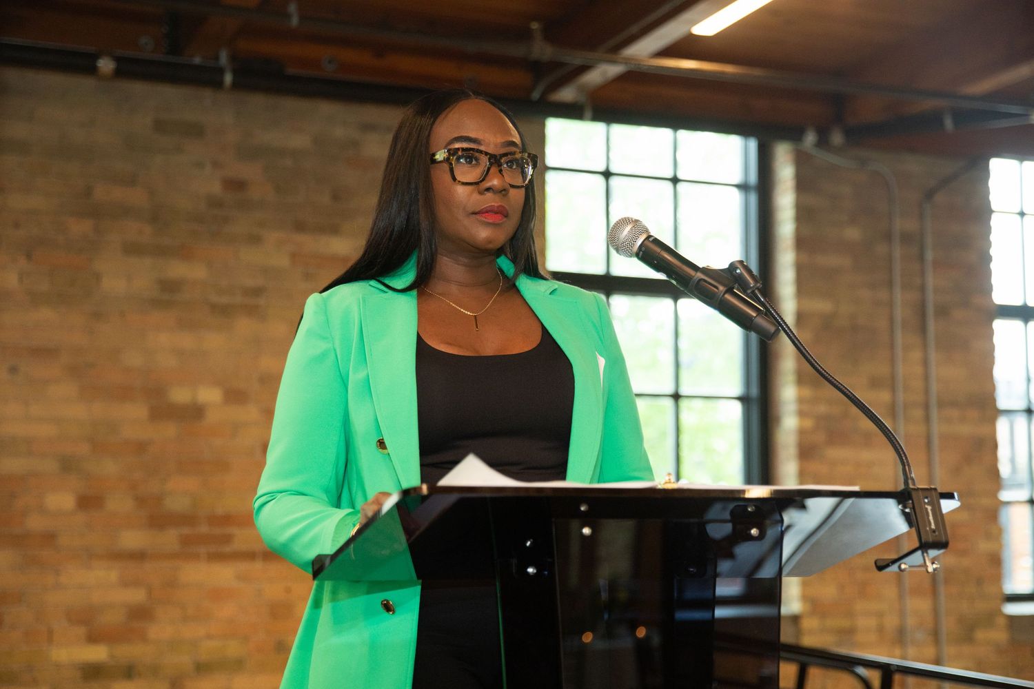 Speaker presents from a podium in a bright green blazer against exposed brick backdrop.