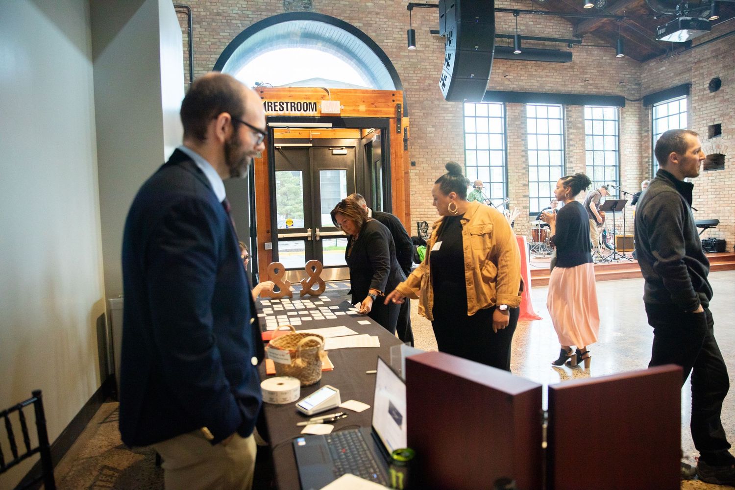 People gathered at an indoor event with brick walls and large windows browse a catering or buffet setup.