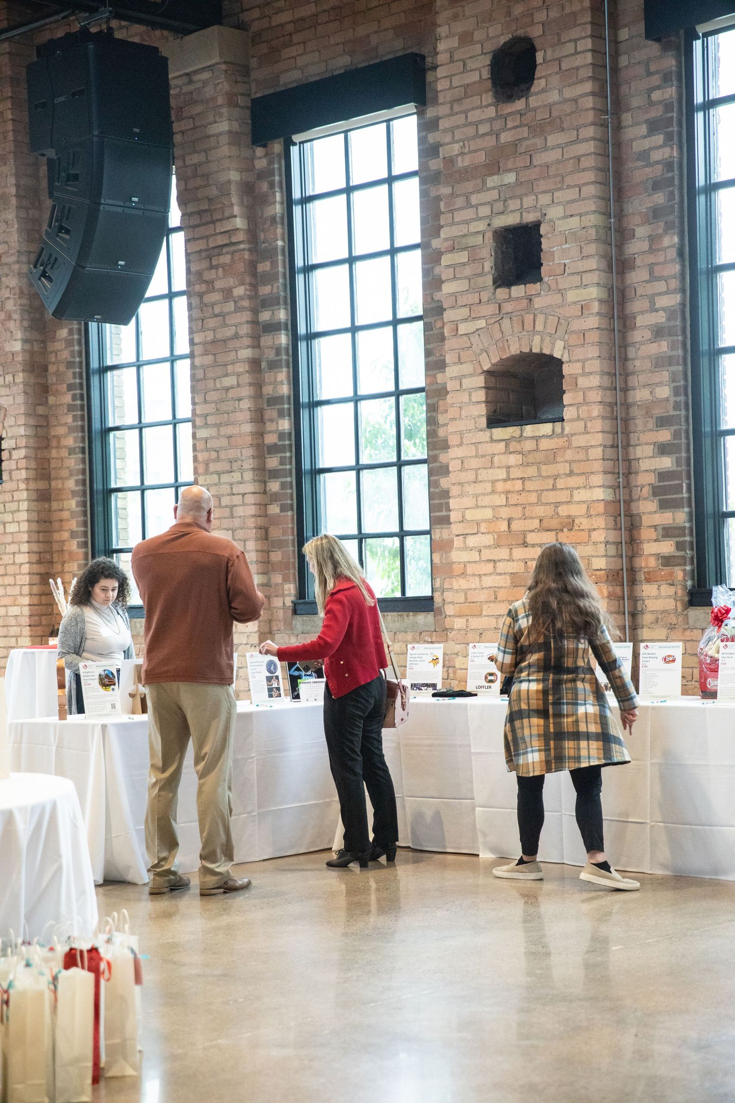 People decorate tables at an event venue with exposed brick walls and tall windows in an industrial-style space.