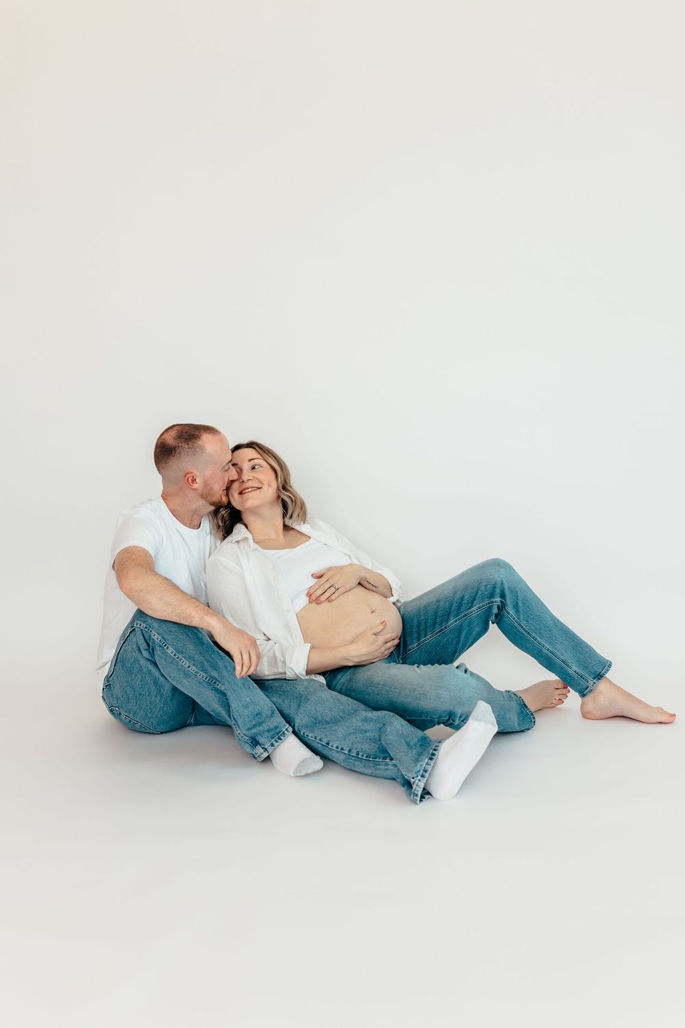 Couple relaxing on floor during maternity photoshoot in minimalist white studio setting. Wilmington NC. Studio session.