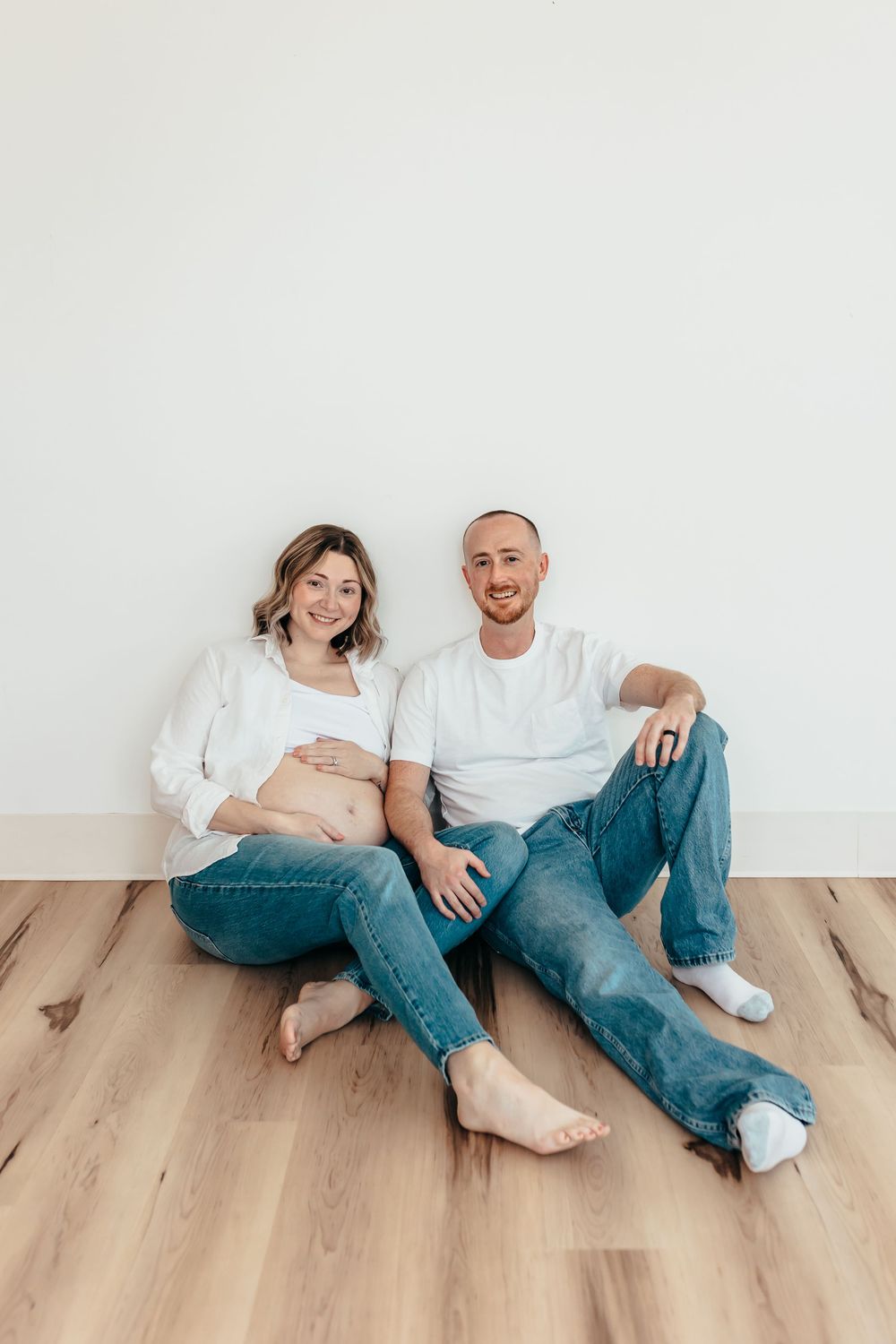 Couple in matching white tops and jeans sitting on wooden floor during maternity session. Wilmington NC. Studio session.