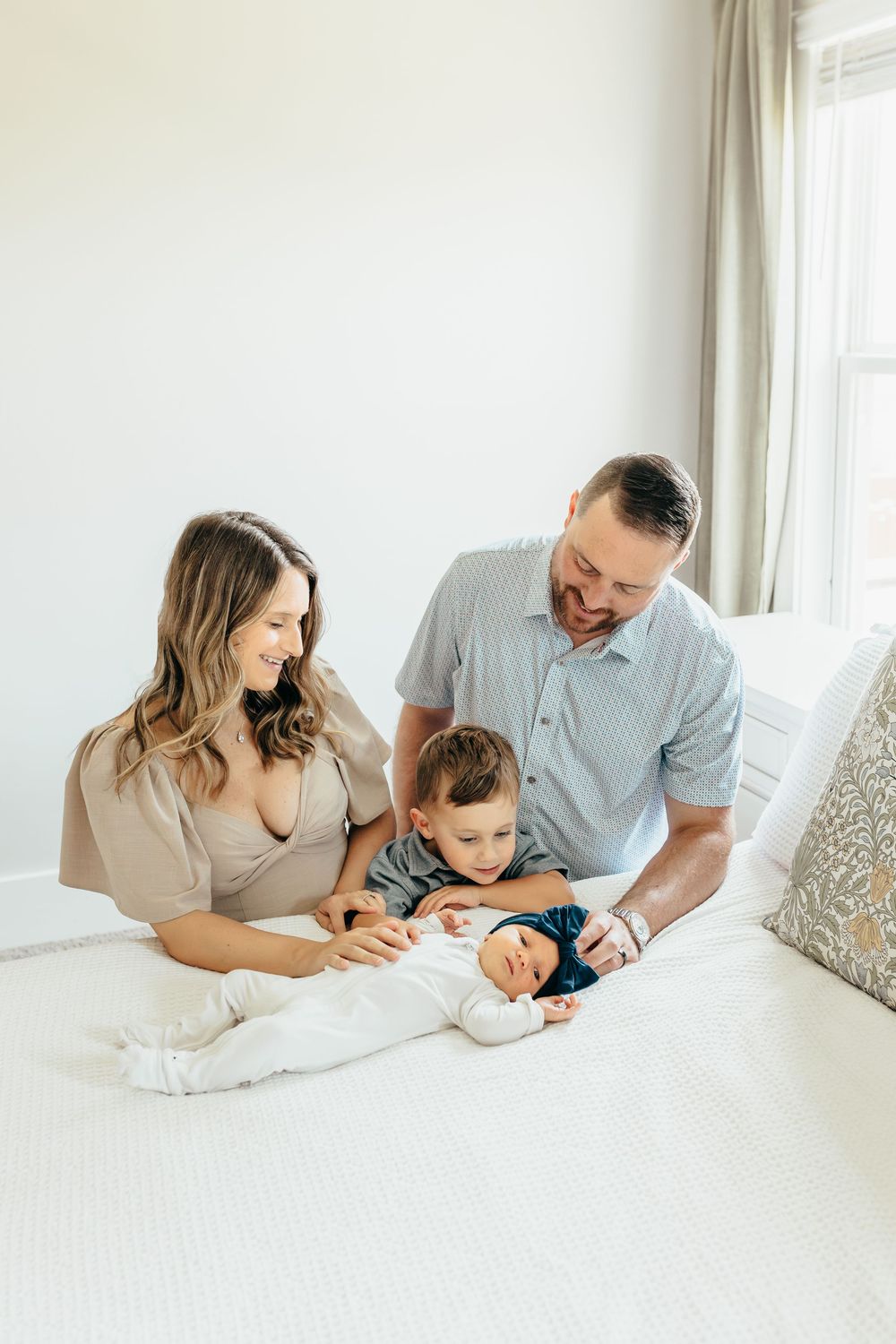 A couple shares a tender moment with their baby while relaxing together on a white bed in a bright room.