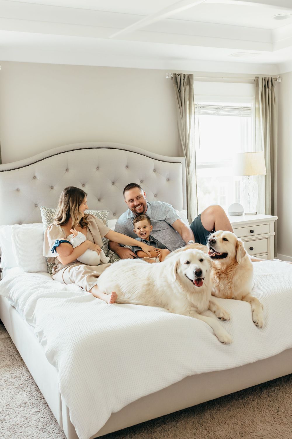 Young family relaxes on white upholstered bed with two golden retriever dogs in bright bedroom.
