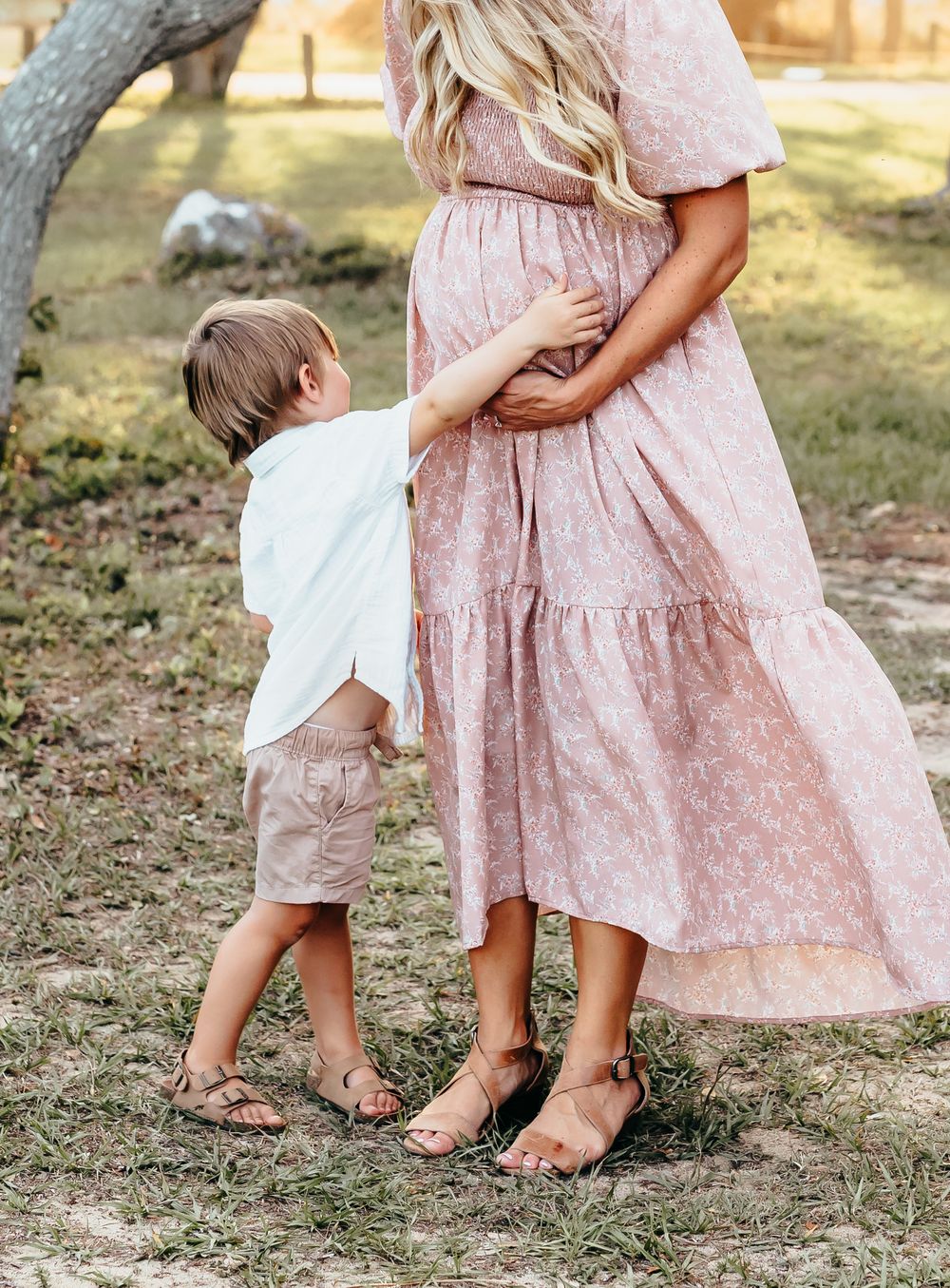 Child hugs parent wearing flowing pink dress outdoors in natural setting.