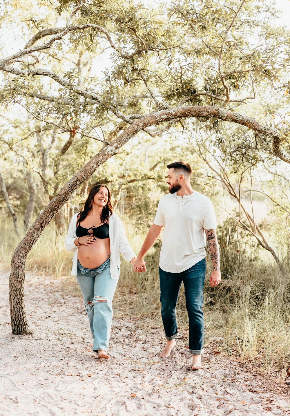 Expectant couple walking hand in hand on a nature trail beneath arching trees.