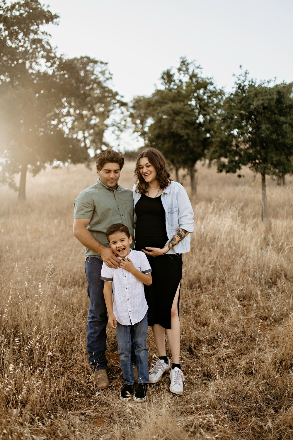 An Amador family of three poses together in a sun-drenched field with golden grass and scattered trees in the background.