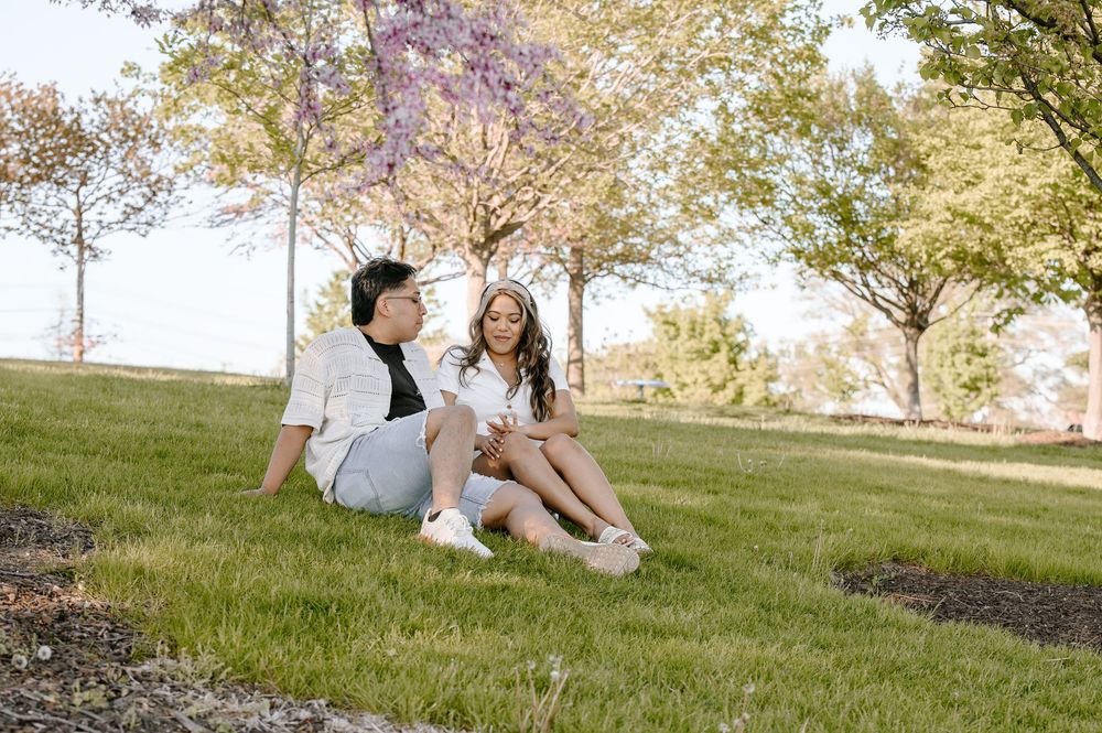 Two people in white summer attire sitting casually on grass under blooming trees in a sunny park setting.