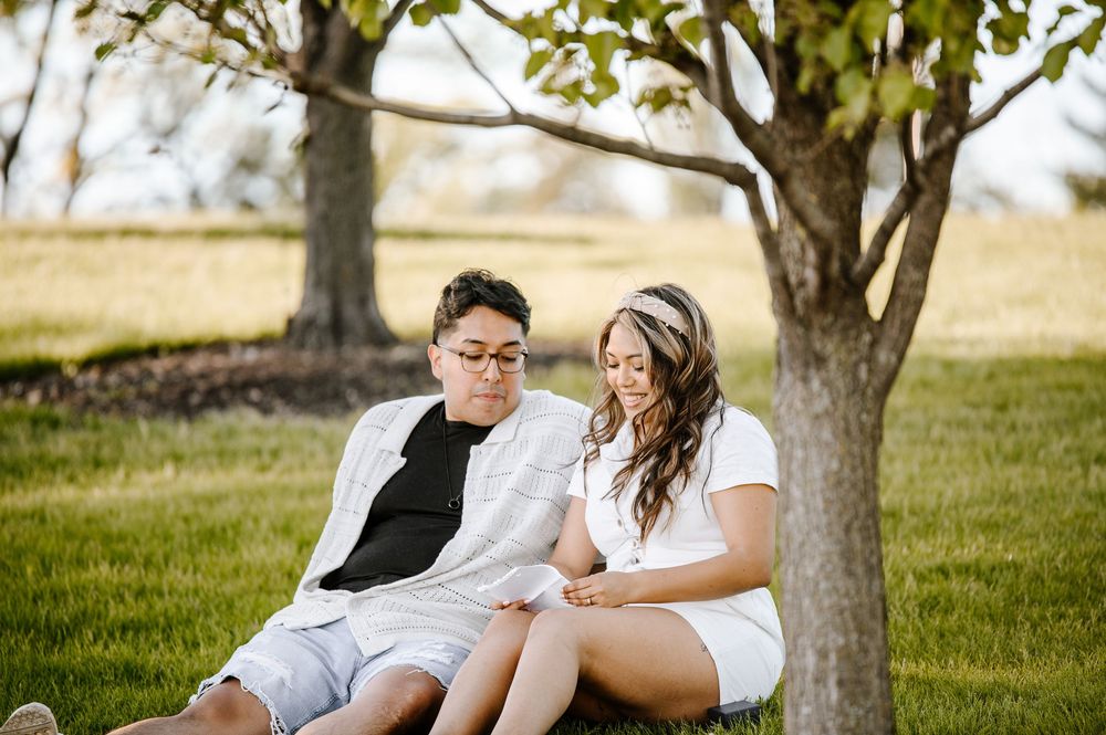 A couple in white outfits share a romantic moment while sitting beneath a tree in a sunlit field.