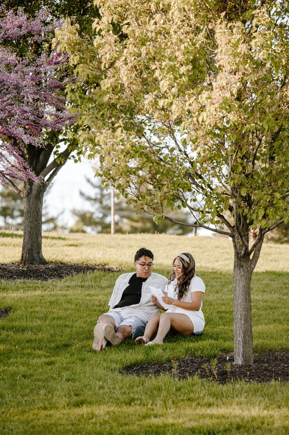A couple sits together under blooming spring trees on a grassy hill during a sunny afternoon.