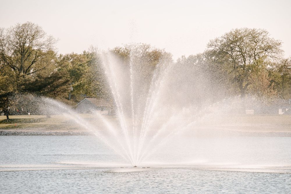 A fountain sprays water in a pattern formation over a tranquil lake surrounded by autumn trees in a park setting.