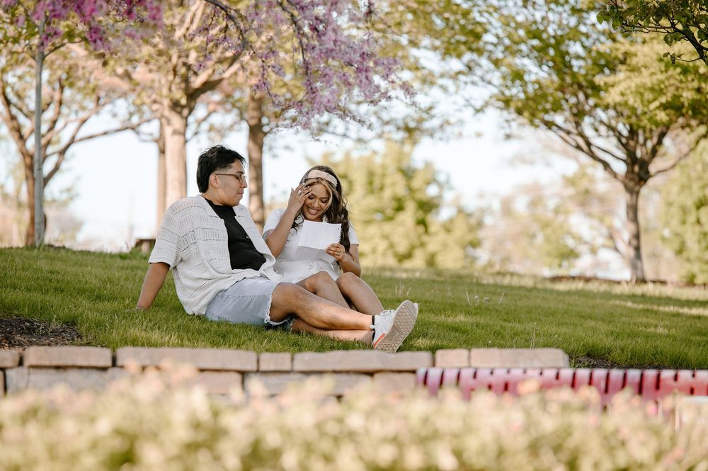 A couple relaxes together on a grassy hill during spring with blooming cherry blossoms in the background.