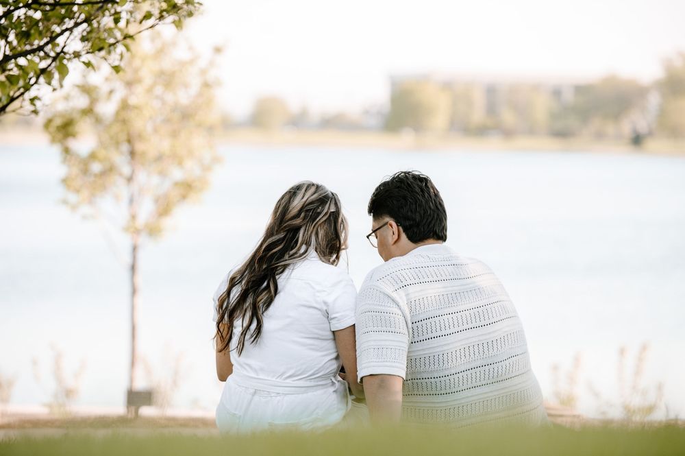 Couple in white outfits sitting together on grass by a serene lake during golden hour.