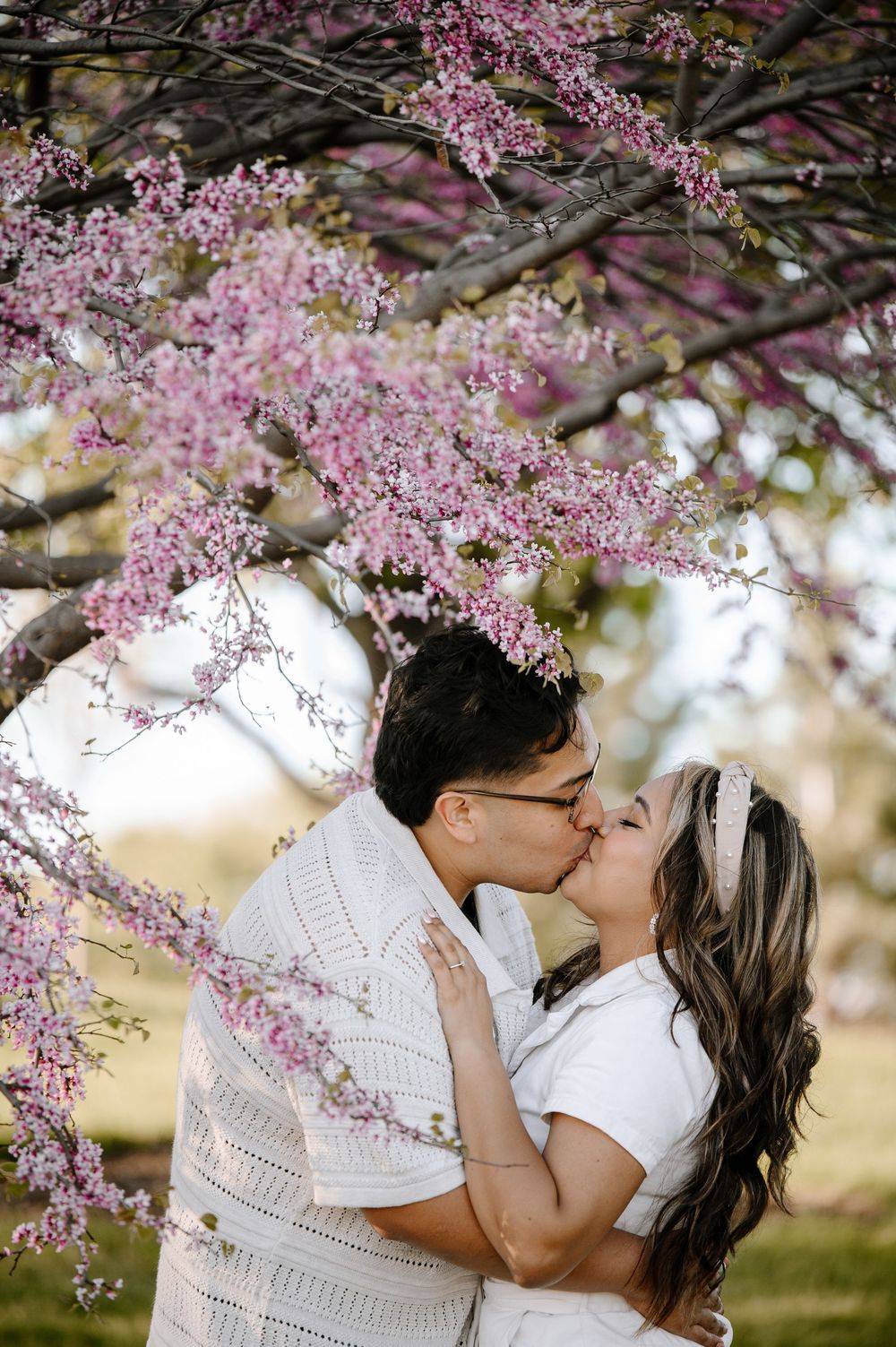 A couple shares a romantic kiss under blooming pink cherry blossoms in a springtime garden setting.