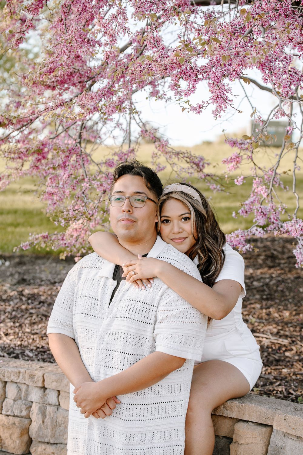 A couple in white outfits poses intimately beneath pink cherry blossoms against a stone wall.